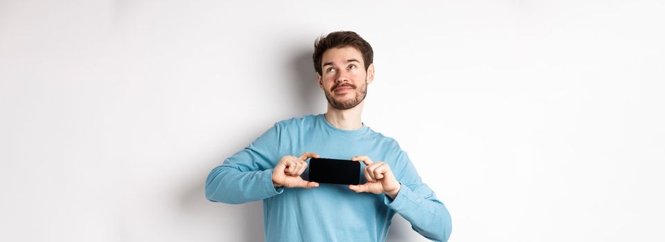 E-commerce and shopping concept. Smiling young man showing mobile screen and dreaming about something, looking at upper right corner nostalgic, standing over white background.