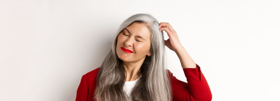 Close up of senior asian woman scratching head and looking bothered by itching, standing over white background.
