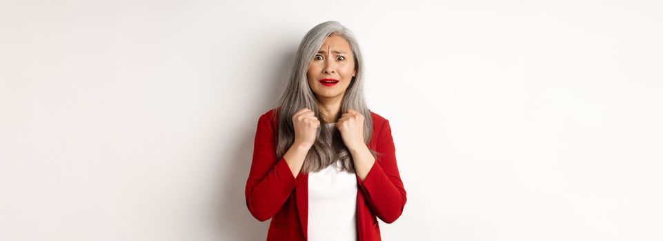 Business people. Scared mature asian woman looking terrified, trembling from fear, standing in red blazer over white background.