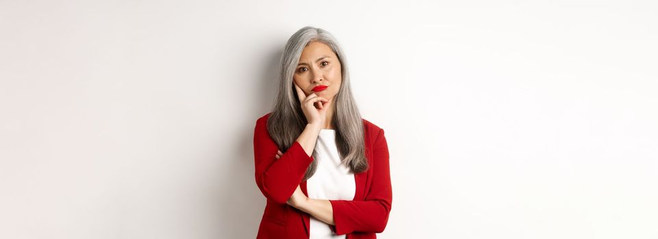 Annoyed and bothered asian businesswoman in red blazer, pouting and looking irritated at camera, standing over white background.