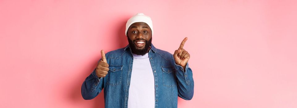 Happy african-american man showing thumb-up and pointing at upper left corner copy space, praising good deal, standing over pink background.