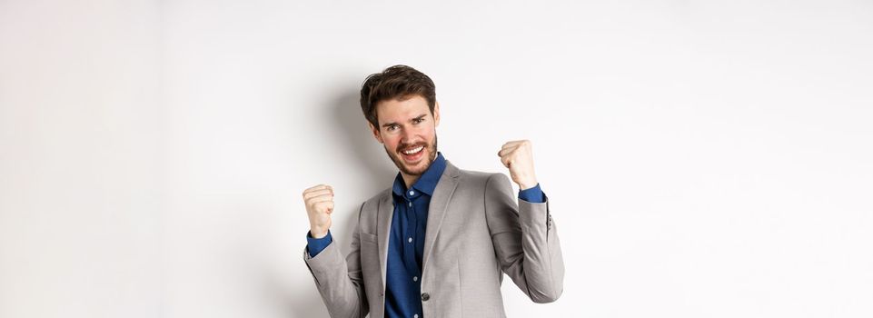 Lucky businessman winning money prize, say yes and smiling excited, make fist pump sign to celebrate victory, triumphing in suit on white background.