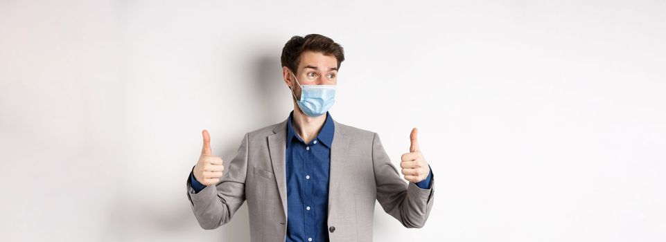Covid-19, pandemic and business concept. Excited businessman in face mask and suit, showing thumbs up and looking aside at logo impressed, praise good thing, white background.