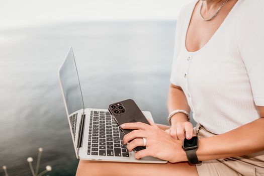 Digital nomad, Business woman working on laptop by the sea. Pretty lady typing on computer by the sea at sunset, makes a business transaction online from a distance. Freelance remote work on vacation