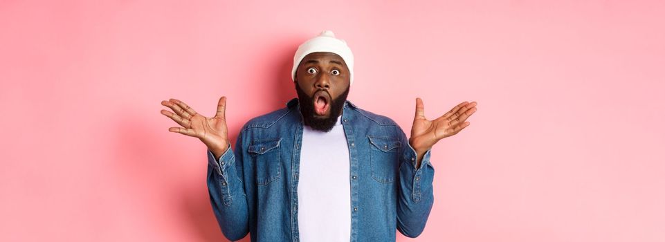 Shocked and impressed Black man staring at camera with complete disbelief, saying wow, standing in beanie and hipster shirt over pink background.
