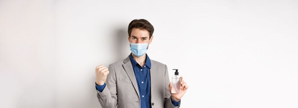 Covid-19, pandemic and business concept. Cheerful office guy in medical mask and suit, looking motivated, showing bottle of antiseptic, white background.