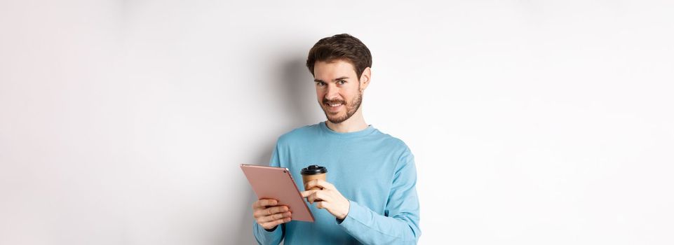 Handsome bearded man smiling at camera, drinking coffee and reading on digital tablet, standing over white background. Copy space