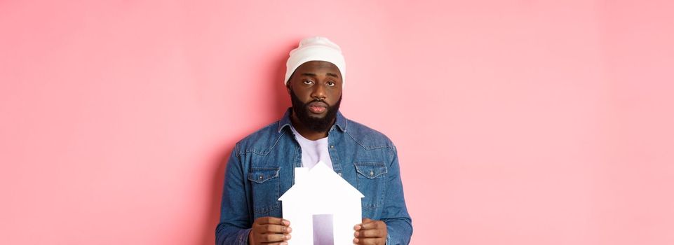 Real estate concept. Sad and tired Black man staring unamused at camera, holding paper house model, standing over pink background.