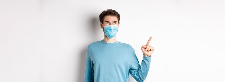 Coronavirus, health and quarantine concept. Man in medical mask smiling pleased, looking and pointing finger left at logo, showing promo offer, white background.