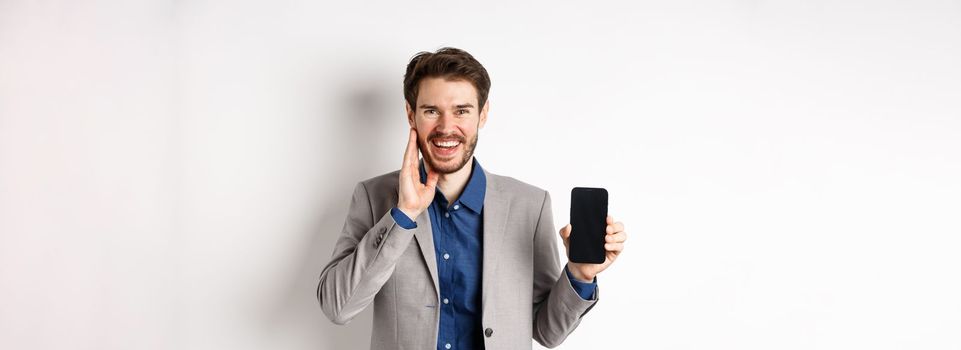 E-commerce and online shopping concept. Happy bearded guy in suit showing empty smartphone screen and laughing, standing on white background.