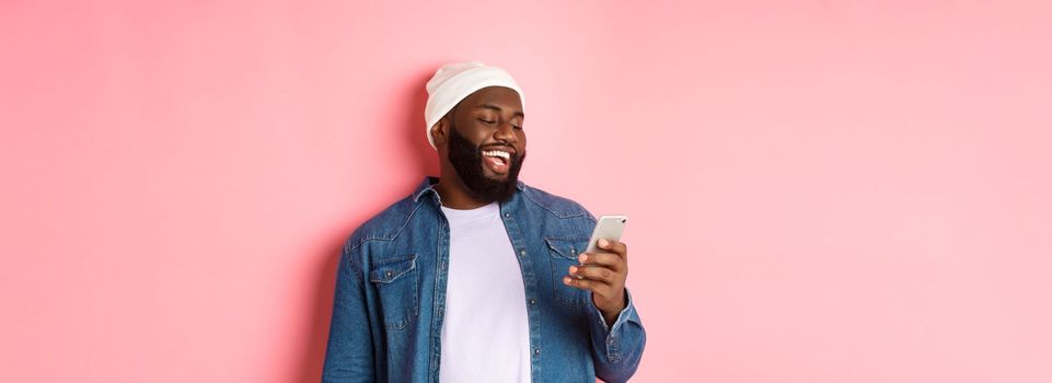 Technology and online shopping concept. Happy Black bearded man reading message and smiling, using smartphone against pink background.