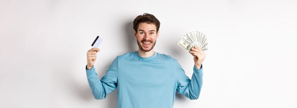 Happy guy smiling and showing plastic credit card with cash, choosing between money and contactless payments, white background.