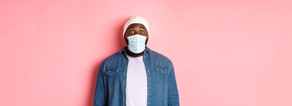 Covid-19, lifestyle and quarantine concept. Image of impressed african-american man in face mask staring at camera, gasping surprised, standing over pink background.