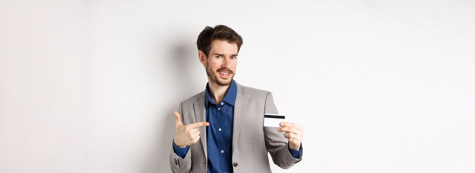 Successful male entrepreneur pointing at plastic credit card and smiling, recommending bank, standing on white background.