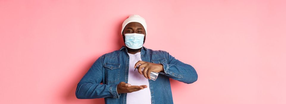 Covid-19, lifestyle and lockdown concept. Smiling african-american man in face mask cleaning hands with sanitizer, using antiseptic and looking at camera, pink background.