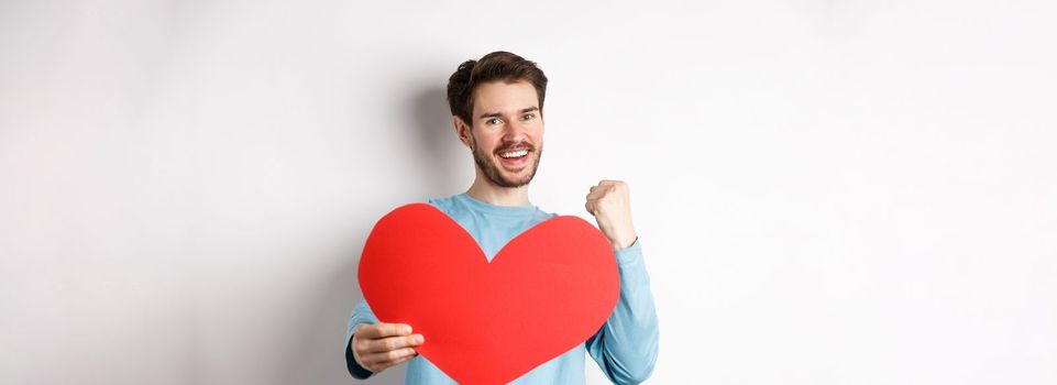 Valentines day. Happy boyfriend triumphing, saying yes and showing valentine red heart, smiling as winning girls love, standing over white background.