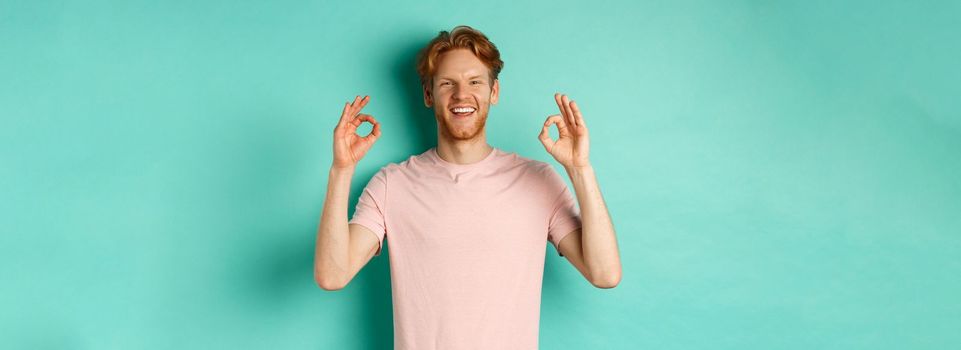 Attractive young man in t-shirt smiling satisfied, nod in approval and showing OK sign, approve and agree with something cool, standing over turquoise background.