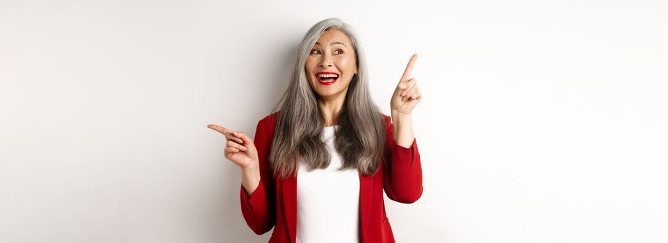 Cheerful asian lady smiling, pointing fingers sideways at two promo offers, standing happy in red blazer over white background.