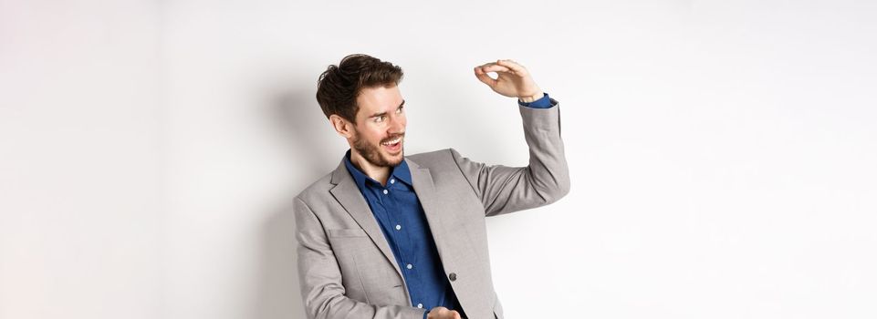 Smiling happy businessman in suit showing large size object, shaping big thing with hands and look pleased, standing on white background.