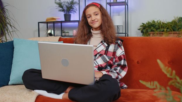 Lovely preteen girl freelancer at home living room sitting on couch, opens laptop start working. Young child, kid works on notebook, sends messages, makes online purchases, watching movies, working