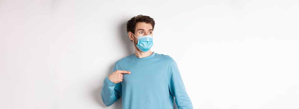 Covid-19, health and quarantine concept. Confused man in medical mask pointing at himself, looking left with ambushed face, standing over white background.