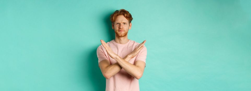 Skeptical redhead man in t-shirt saying no, cross arms on chest and frowning disappointed, prohibit something bad, standing over turquoise background.