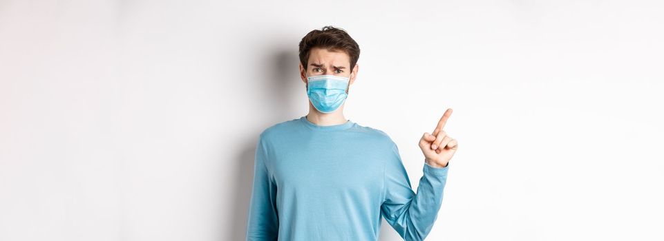 Coronavirus, health and quarantine concept. Skeptical man in medical mask pointing finger left at logo, frowning disappointed, standing over white background.