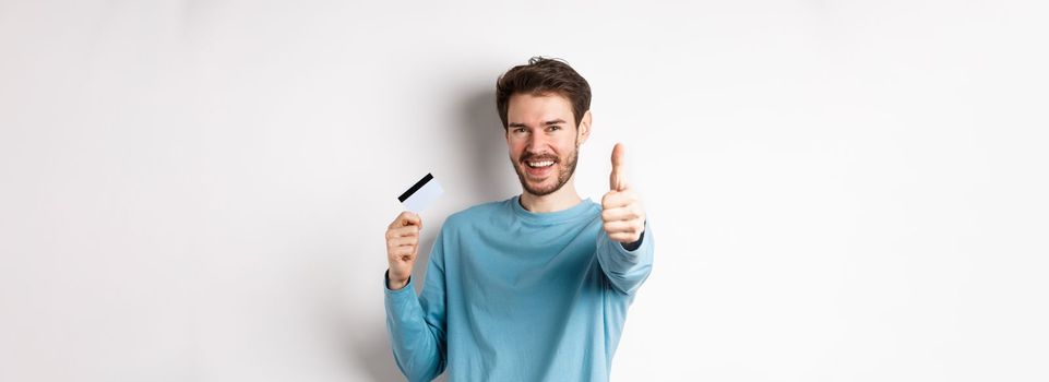 Cheerful young man showing plastic credit card and thumb up, like and approve good bank service, standing over white background.