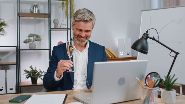Mature businessman working on laptop celebrate successful contract agreement with colleague client at office. Freelancer middle aged man drinking champagne. Online remote video call conversation job
