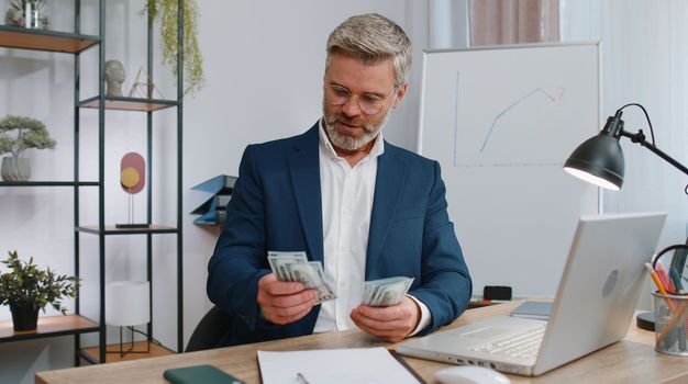 Rich mature businessman working on laptop pc counting money cash, calculate earnings income profit at office workplace. Professional manager freelancer man. Business old people. Employment, occupation