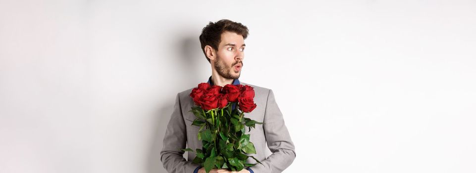 Nervous boyfriend waiting for his date on valentines day, looking left amazed, holding bouquet of red roses, standing in suit over white background.