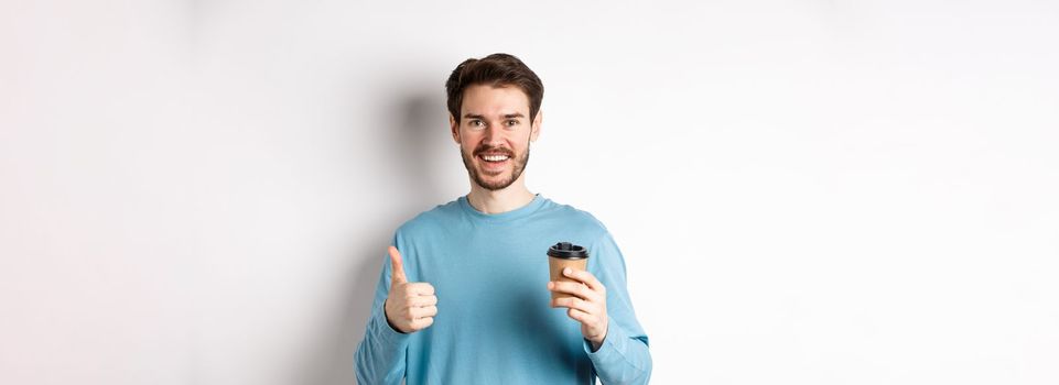 Cheerful young man drinking good coffee, holding paper cup and showing thumb up, recommending cafe shop, standing over white background.