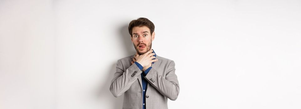 Distressed young man in business suit strangle himself and showing tongue, feeling sick with sore throat, standing on white background.