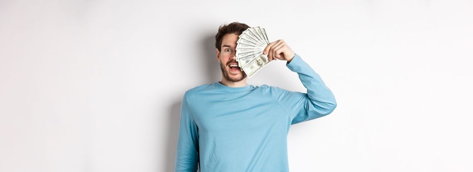 Surprised and happy young man covering half of face with money, gasping amazed at camera, checking out quick loan offers, standing over white background.