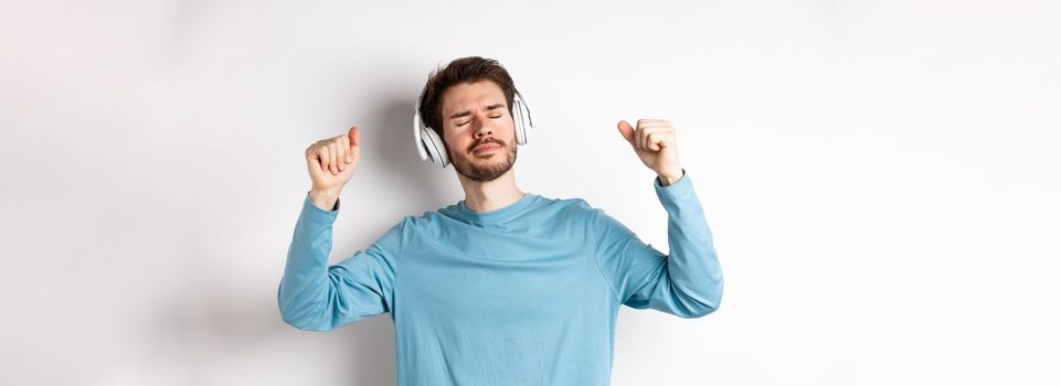 Happy handsome man dancing to music in headphones, listening songs and smiling, standing over white background.