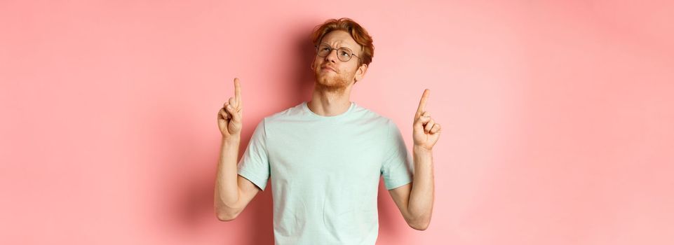 Doubtful redhead guy in glasses frowning, looking and pointing fingers up at something disappointed, standing over pink background.