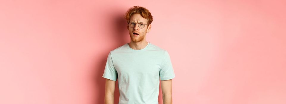 Portrait of handsome redhead man in glasses looking confused, open mouth and stare at camera, cant understand nothing, standing over pink background.