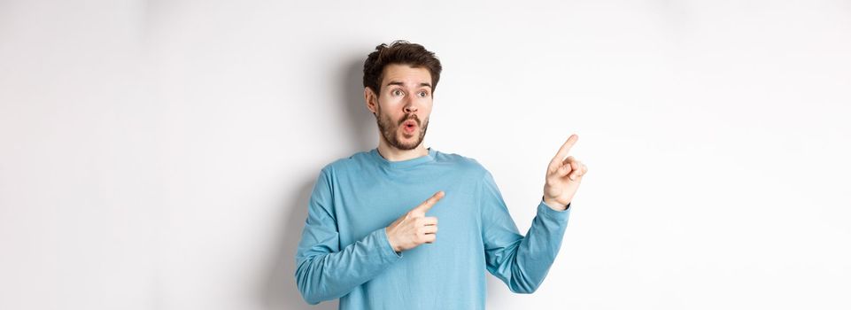 Wow check it out. Impressed young man pointing and looking left at special promo offer or discounts on white background, gasping amazed, standing in studio.