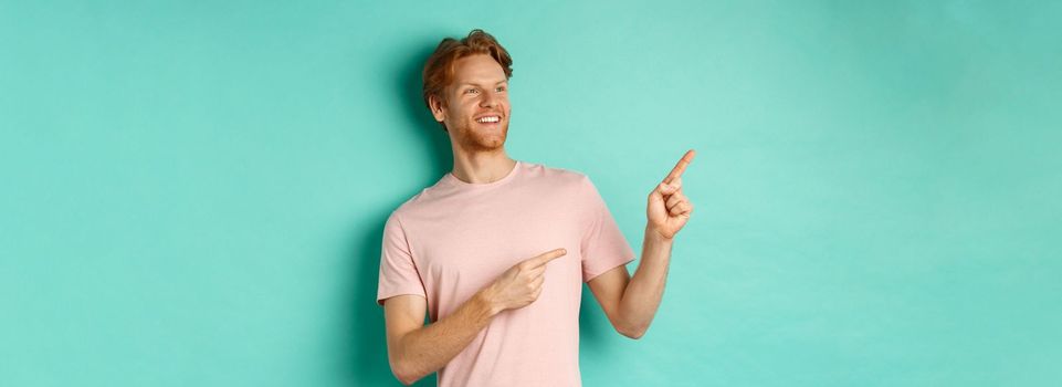 Handsome smiling man with red hair and beard looking delighted, pointing at upper left corner banner, standing in t-shirt over mint background.