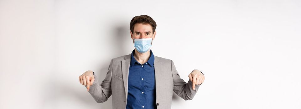 Covid-19, pandemic and business concept. Handsome businessman in medical mask and suit, pointing fingers down and showing company logo, white background.