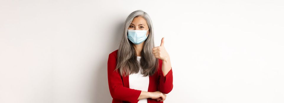 Coronavirus and business concept. Asian female entrepreneur in face mask looking cheerful, showing thumb-up in approval, praising using protective measures at work.