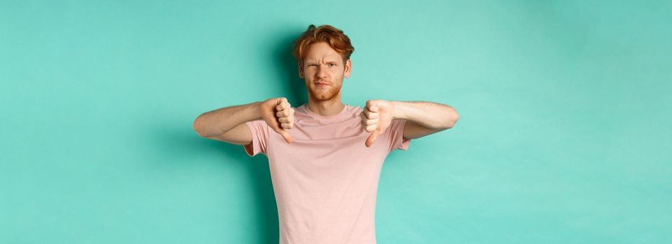 Disappointed guy with red hair showing thumbs-down, frowning and looking skeptical, epxress dislike and disapproval, standing over turquoise background.