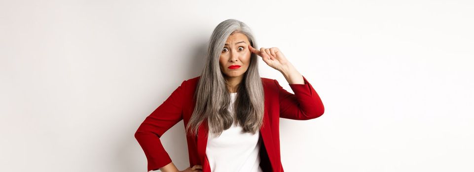 Annoyed asian business woman scolding someone stupid, showing crazy gesture, pointing finger at head and staring at camera, white background.