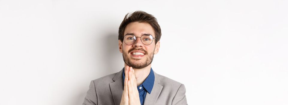 Close-up of happy thankful guy in glasses and suit showing gratitude, make namaste sign and smiling, standing on white background.