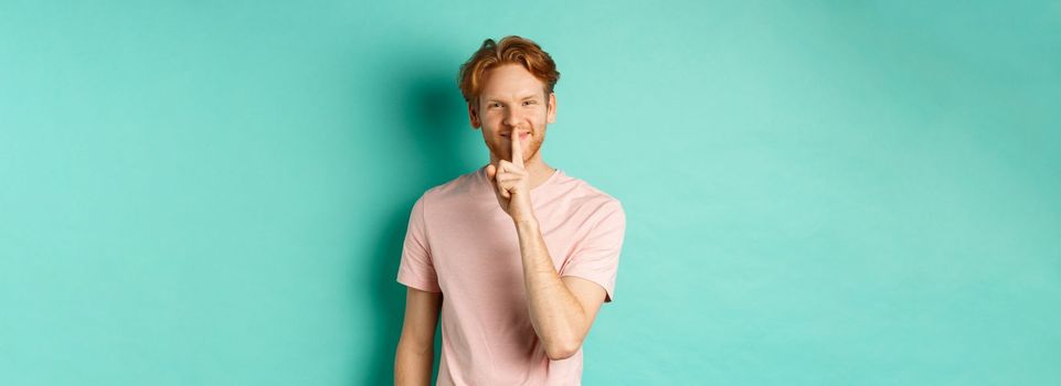 Smiling young man with red hair and beard sharing a secret, showing taboo gesture and grinning, shushing you to be quiet, standing over turquoise background.