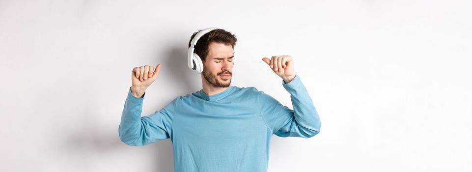 Happy young man having fun in headphones, dancing while listening music in wireless earphones, white background.