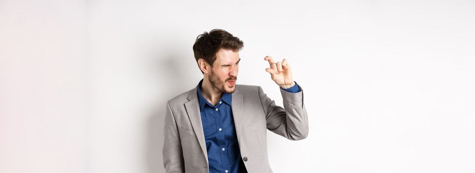 Businessman trying to see something tiny, showing little thing size with fingers, standing in suit on white background.