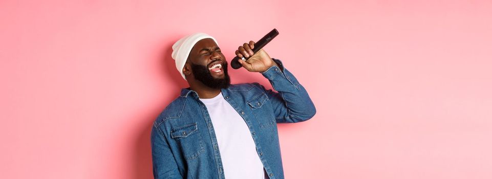 Handsome Black man in beanie and denim shirt singing karaoke, holding microphone, standing over pink background.