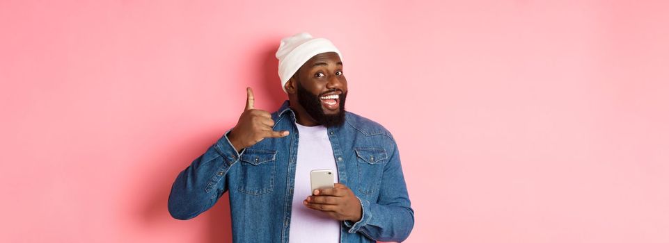 Happy Black man showing call me sign, making phone gesture and smiling, holding smartphone, standing in beanie and denim shirt over pink background.