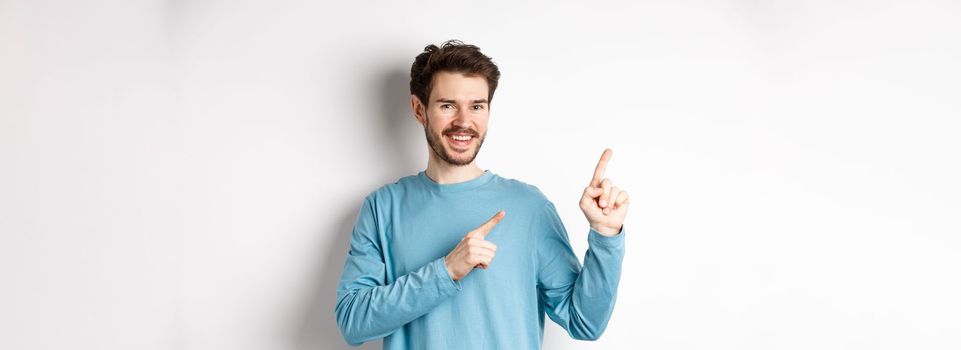 Cheerful bearded man showing advertisement on copy space, pointing fingers at upper left corner logo and smiling, standing over white background.